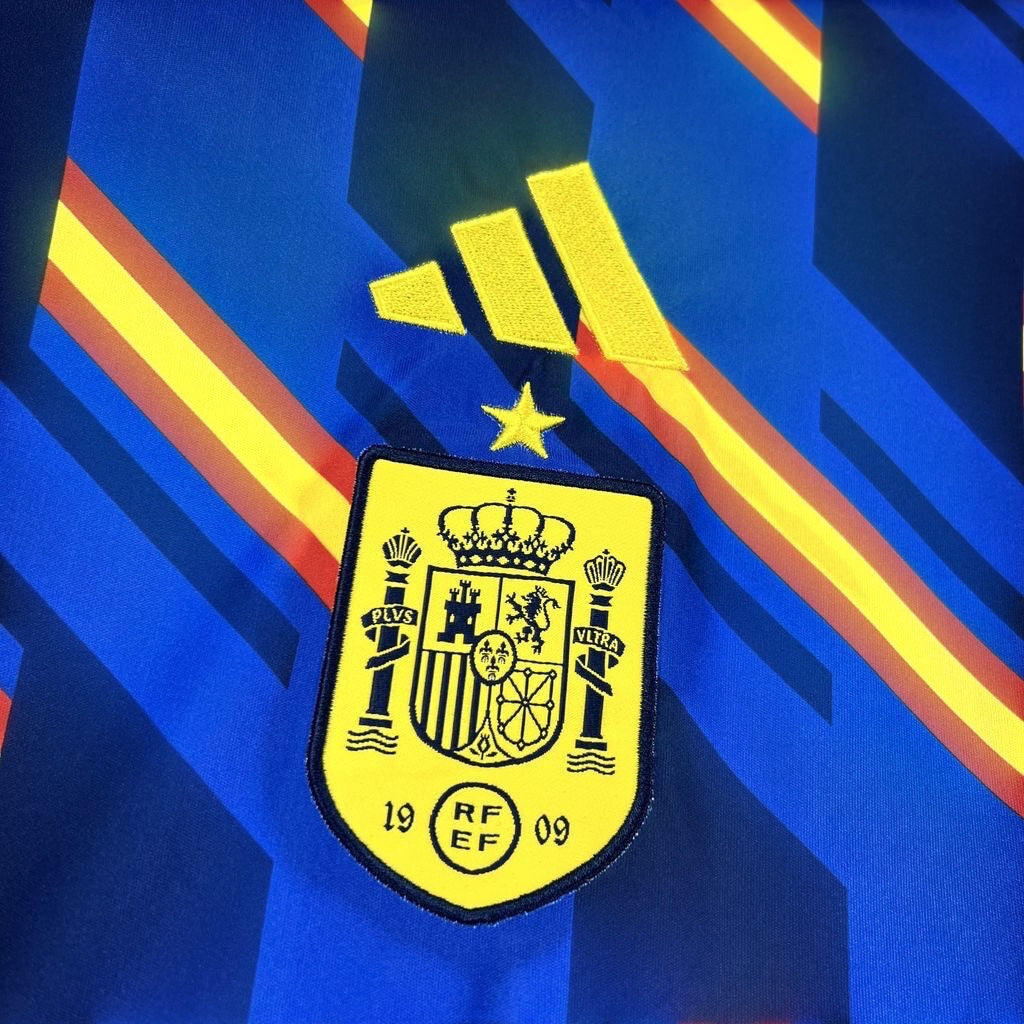Leazofoot Spain pre-match football jersey 2026, blue with yellow and red diagonal stripes, featuring the Adidas logo and the Spanish crest.