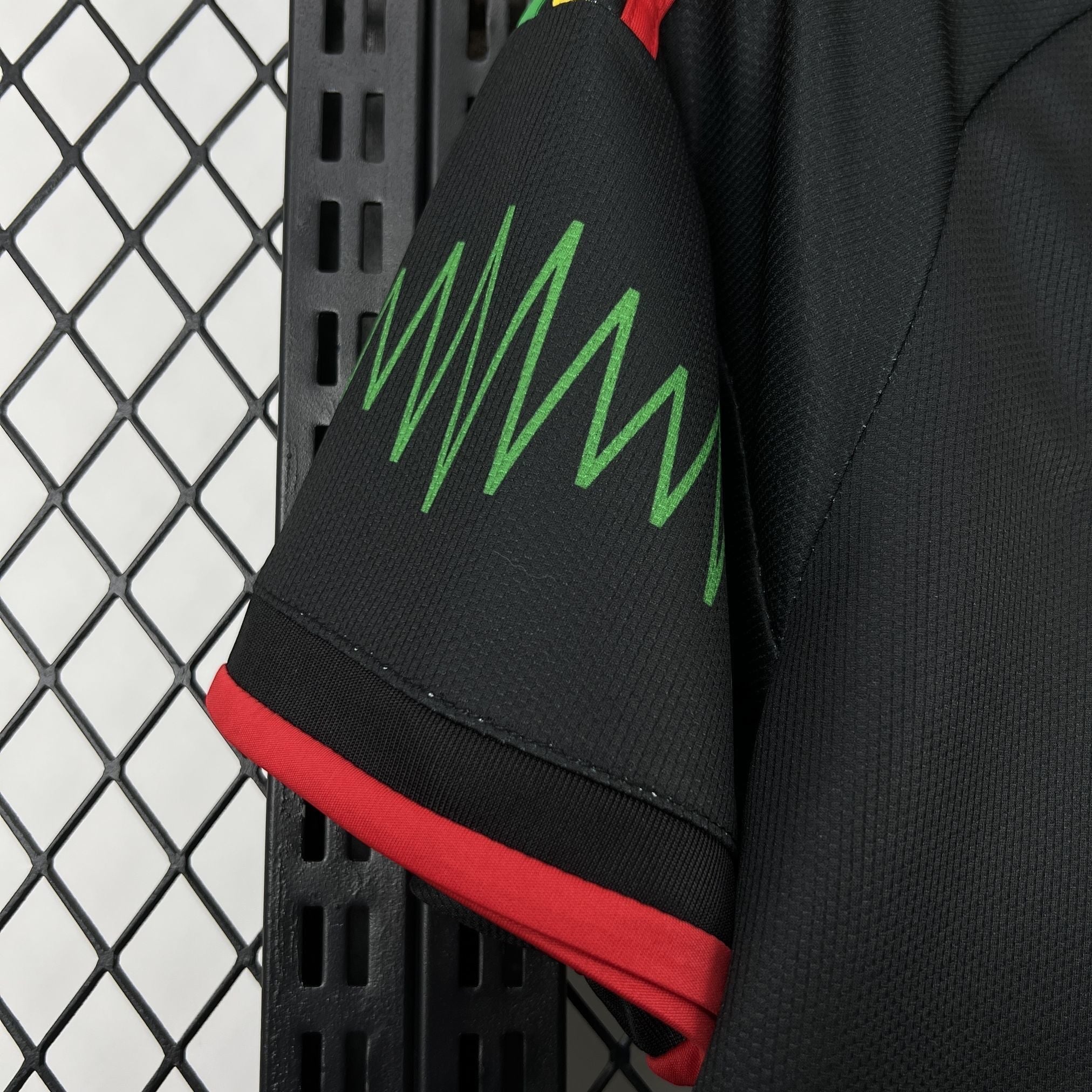 Leazofoot Jamaica 2026 away jersey sleeve detail featuring black fabric with green zigzag pattern and red trim.