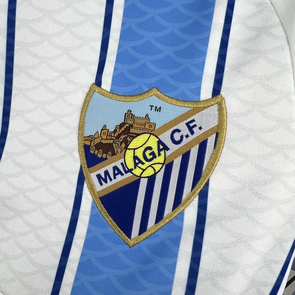 Leazofoot Malaga CF 26/27 home jersey featuring a detailed embroidered club crest on white and blue striped fabric.